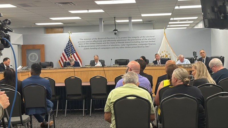 A board meeting in progress with officials seated at a long table and an audience observing.