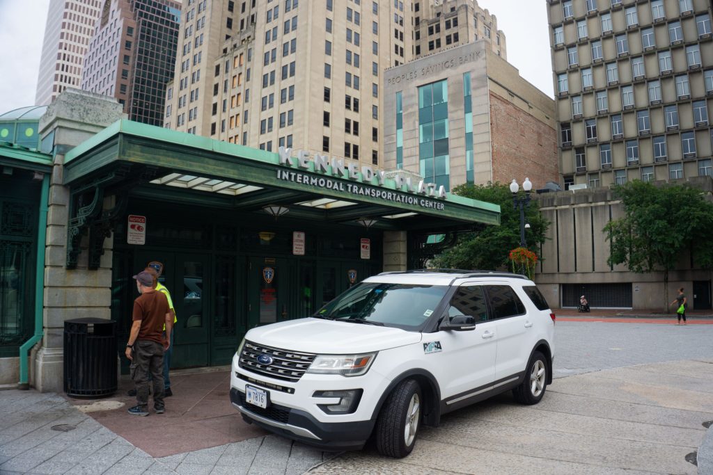 Two men converse near the entrance of Kennedy Plaza Intermodal Transportation Center, with a white vehicle parked nearby.