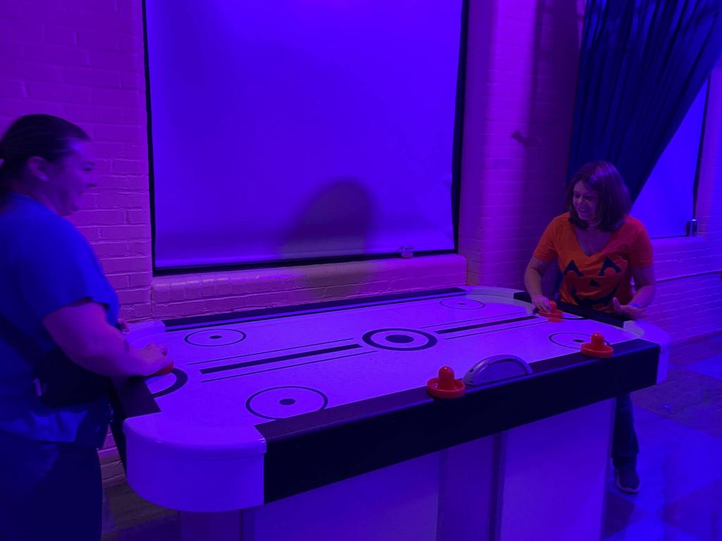 Two women play air hockey in a dimly lit room with purple lighting. One wears a pumpkin-themed shirt.