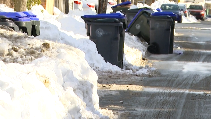 Snow-covered street with several trash bins lined up along the curb.