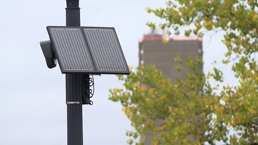 A solar-powered camera mounted on a pole, with trees and a building in the background.