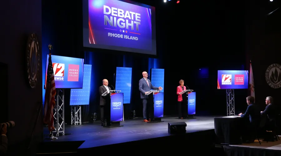 Three candidates stand at podiums during a debate night event in Rhode Island, with an audience visible in the foreground.