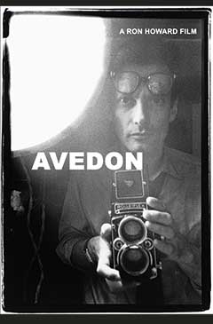 A man holds a vintage camera, wearing glasses, with a bright light source in the background.