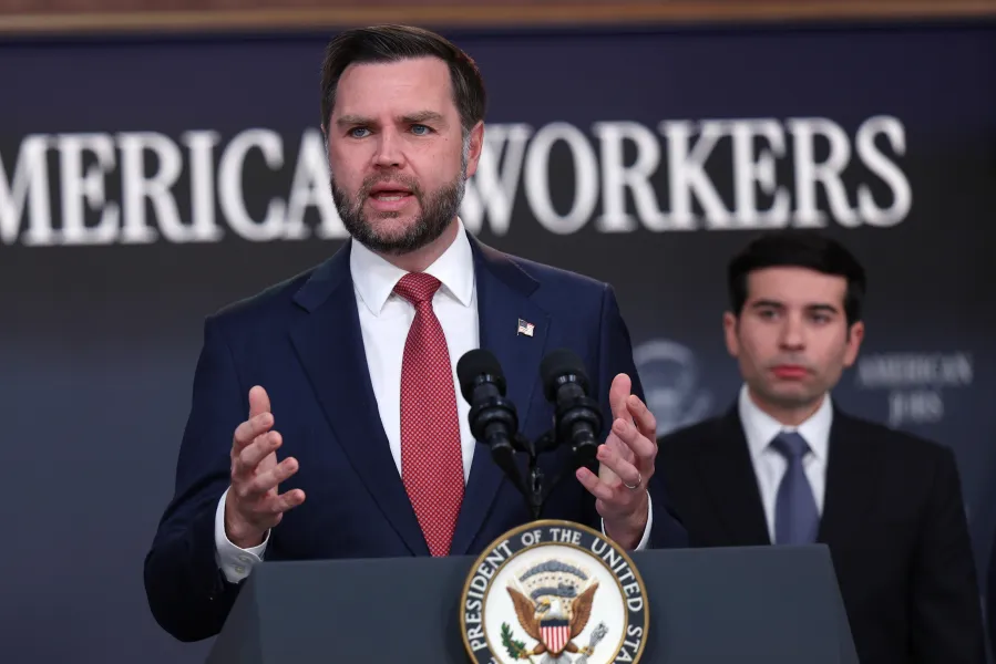 A man in a suit speaks at a podium with a presidential seal, while another man listens in the background.