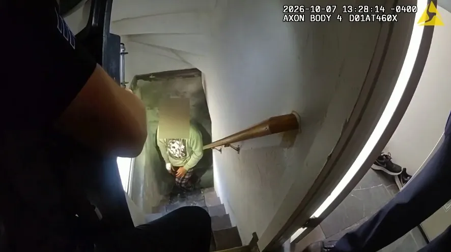 A person in a green shirt descends stairs, hands behind their back, while an officer observes from above.