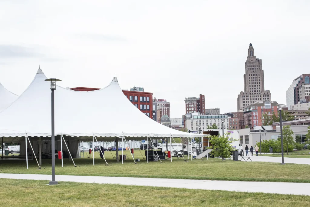 A large white tent stands on grass, with city buildings in the background and people walking nearby.