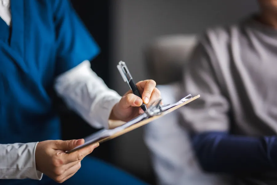 A healthcare professional writes notes on a clipboard while speaking with a patient.