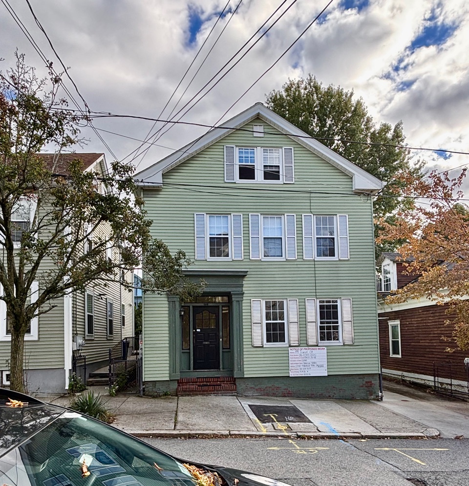 Green two-story house with white shutters, trees nearby, and a sign on the front. Power lines overhead.