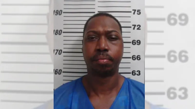Mugshot of a man in a blue jumpsuit, standing against a height chart.