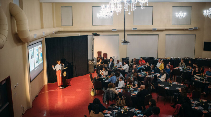 A speaker presents to an audience seated at tables in a banquet hall.
