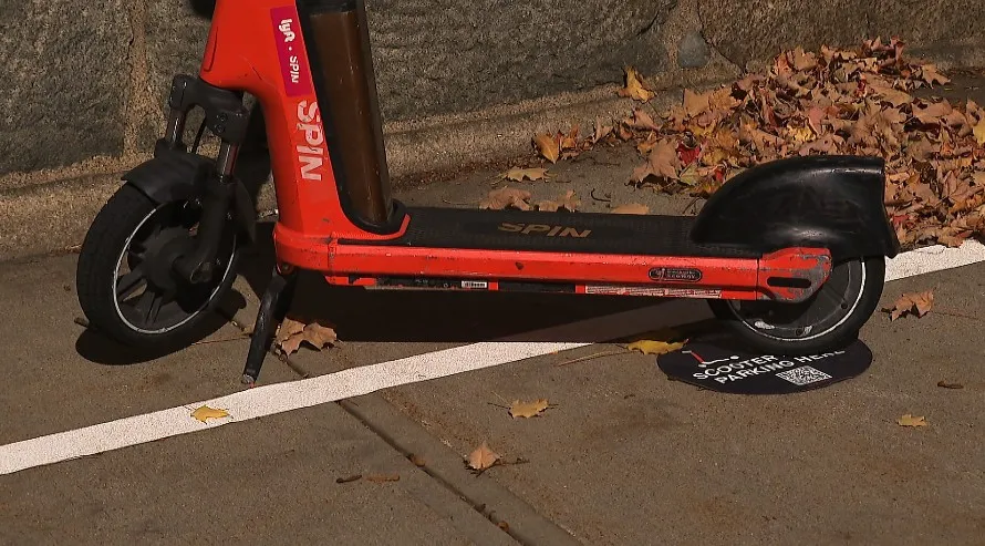 A red Spin scooter parked beside a sign indicating scooter parking rules, surrounded by fallen leaves.