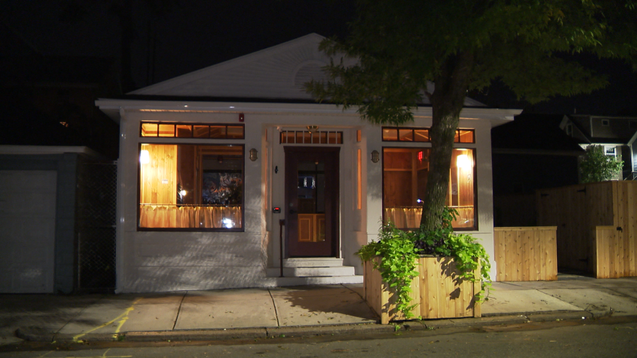 A well-lit house at night with wooden accents, surrounded by greenery and wooden fences.