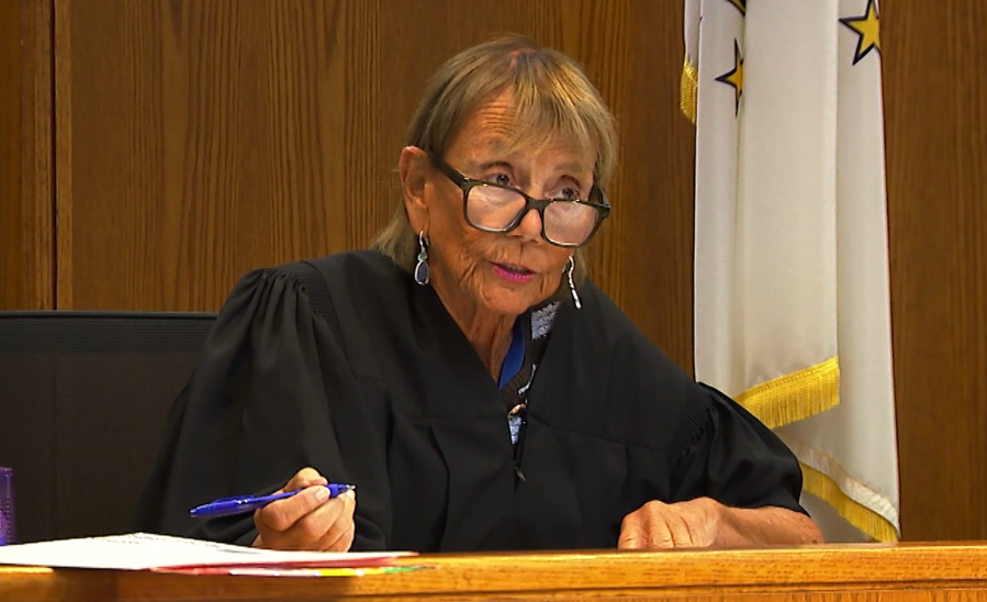 A judge in a black robe speaks while holding a pen, with flags in the background.