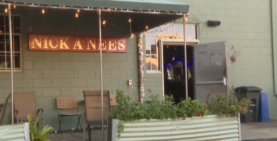 Exterior of Nick A Nees bar with a green wall, open door, and seating area. Plants in a planter box.