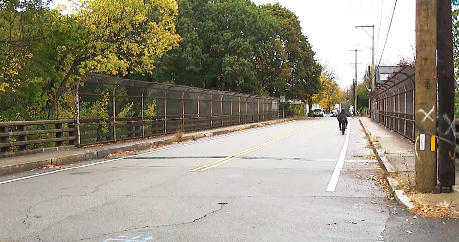 A cyclist rides on a quiet street beside a fenced bridge, surrounded by autumn trees.