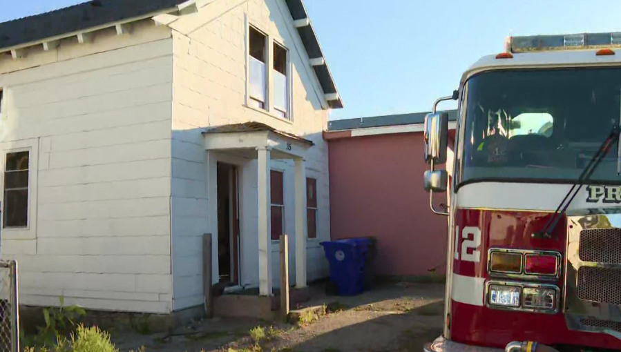 Fire truck parked near a white house with open front door and blue trash bins.