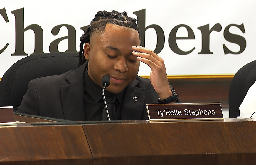 A young man in a suit sits at a table, resting his forehead on his hand, appearing contemplative.