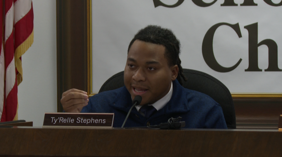 Ty'Relle Stephens speaks at a meeting, gesturing with one hand, seated at a desk with a nameplate.