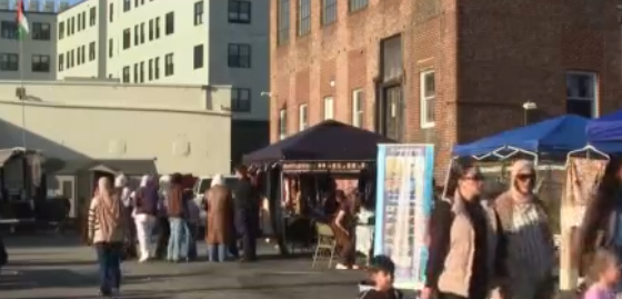 People walk through a market with tents and displays near a brick building.