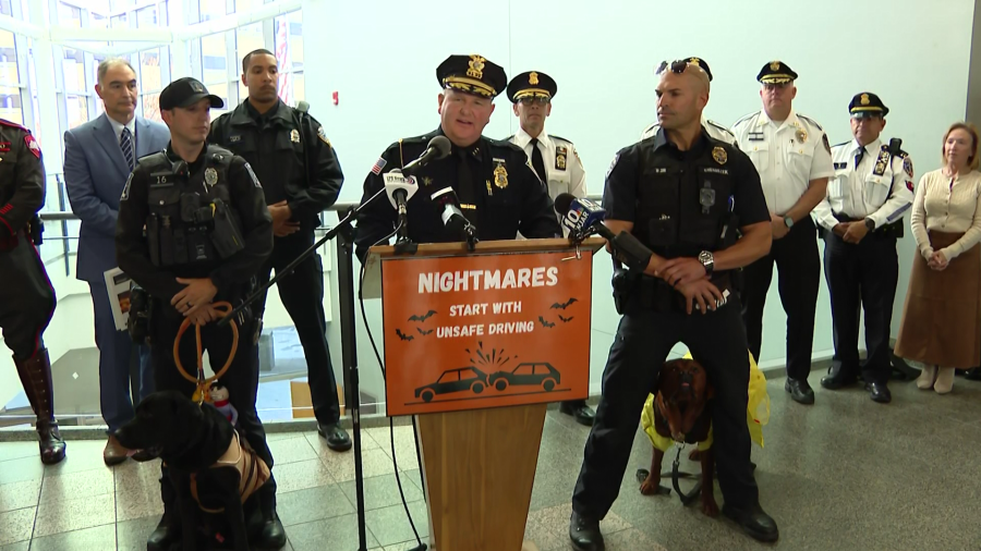 A police chief speaks at a podium about safe driving, flanked by officers and service dogs.