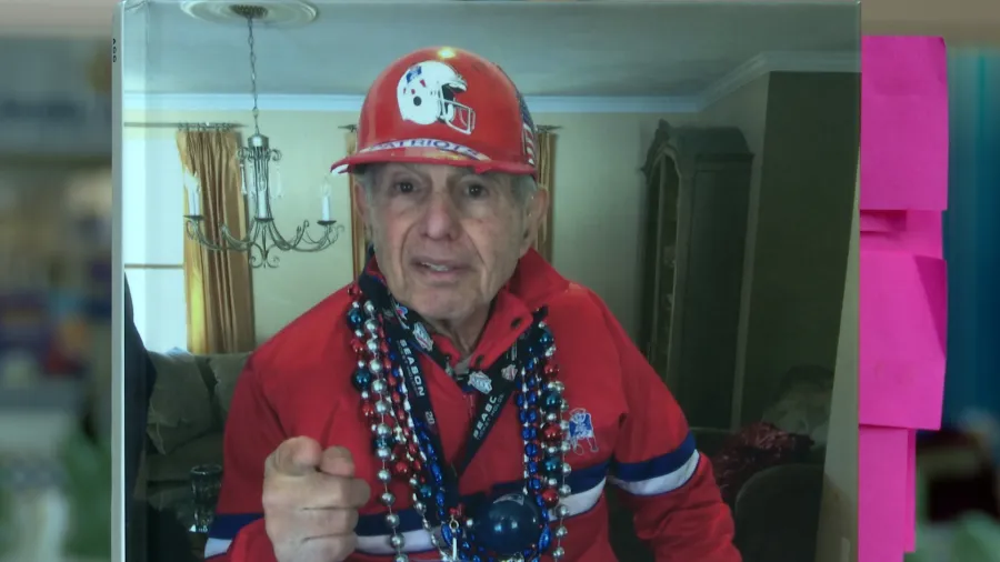 An elderly man in a red sports outfit and hat gestures while wearing colorful beads.