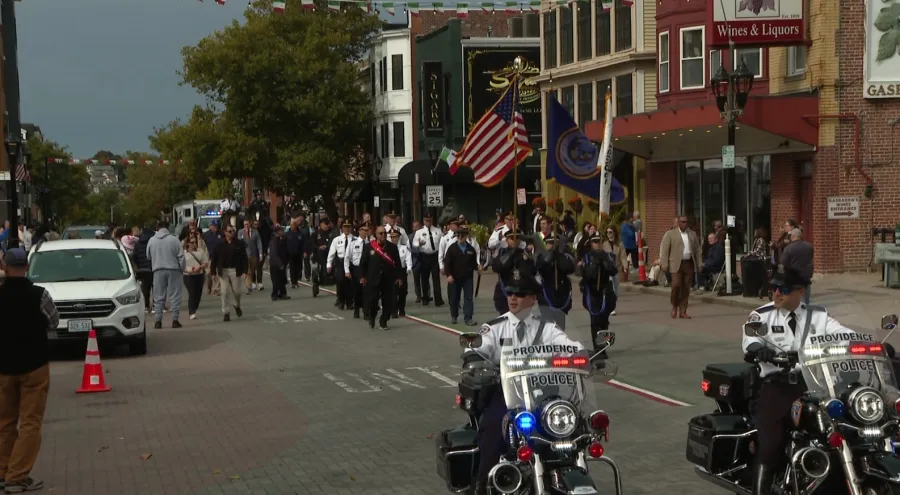 Police officers on motorcycles lead a parade with flags and a crowd walking along the street.