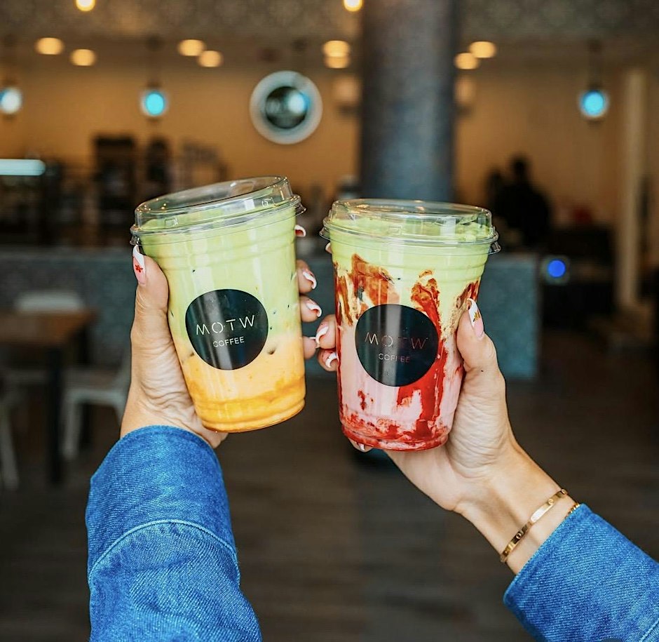 Two hands hold colorful drinks in clear cups with a black logo, set against a café background.