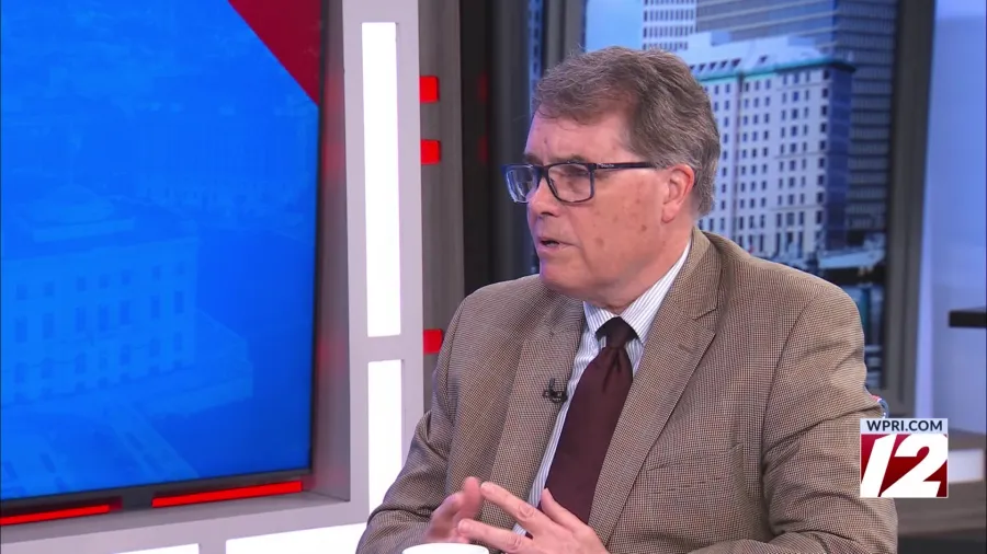 A man in a suit speaks thoughtfully during a televised interview, with a cityscape in the background.