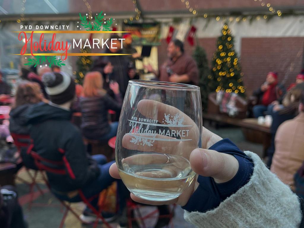 A hand holds a glass with "Holiday Market" text, while people gather and enjoy the festive atmosphere.