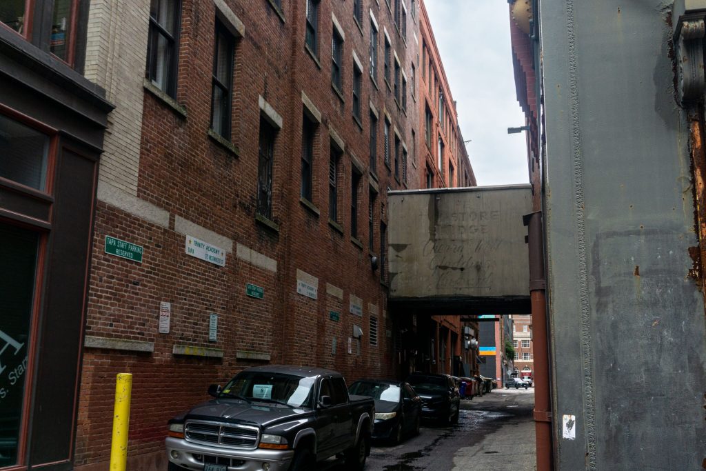 Narrow alleyway between brick buildings with parked cars and a faded overhead sign.